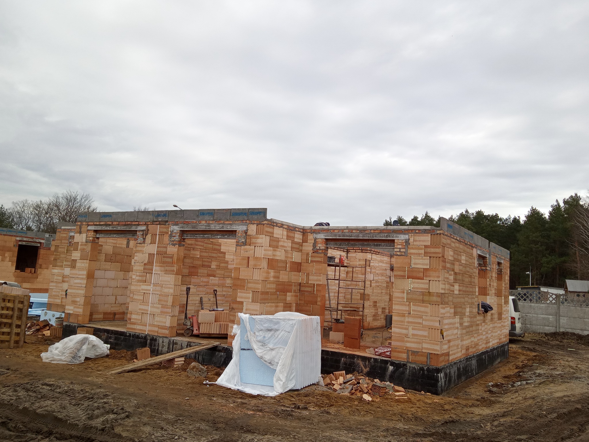 Wznoszenie ścian parterowego domu z czerwonej cegły. Widoczne narzędzia, rusztowanie i materiały budowlane. Fundamenty zabezpieczone. Pochmurne niebo w tle.