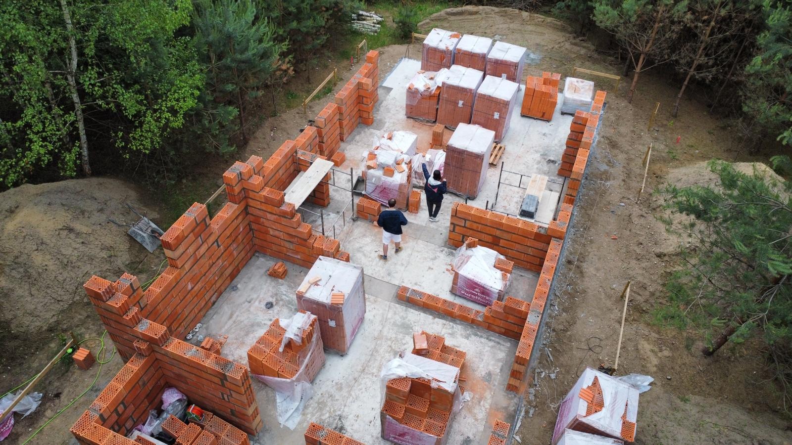 Budowa domu z czerwonej cegły ceramicznej. Widok z góry na fundamenty i wznoszone ściany. Na placu budowy palety z cegłami i dwie osoby. Otoczenie: las.