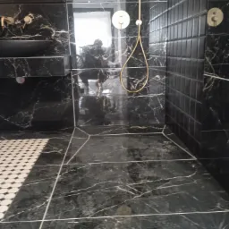 Nowoczesna łazienka z czarnymi płytkami imitującymi marmur, złotą armaturą prysznicową i fragmentem podłogi z biało-czarną mozaiką.