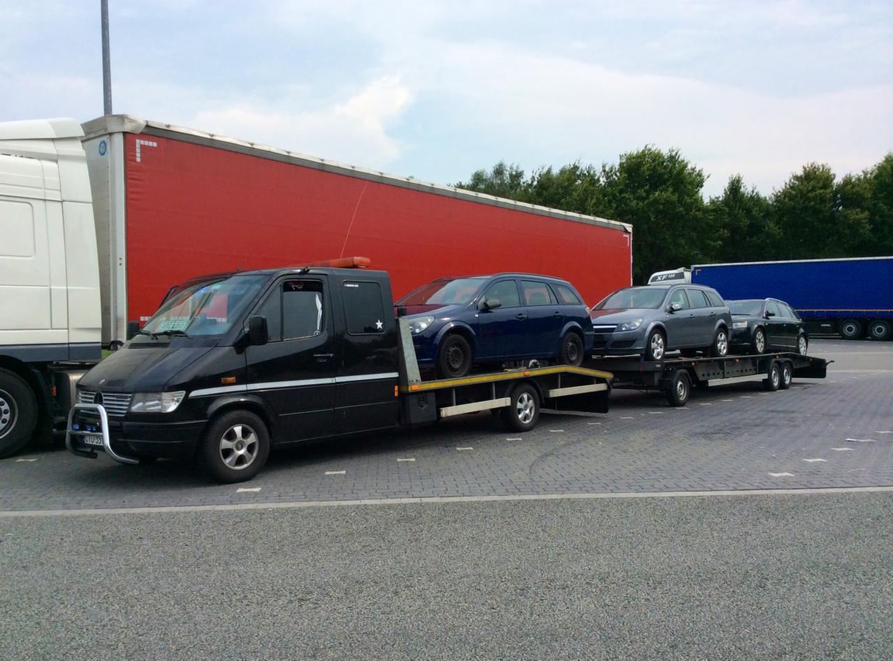 Czarna laweta z trzema samochodami osobowymi marki Opel Astra kombi, zaparkowana na parkingu z widocznymi naczepami tirów w tle. Pomarańczowe światło ostrzegawcze.
