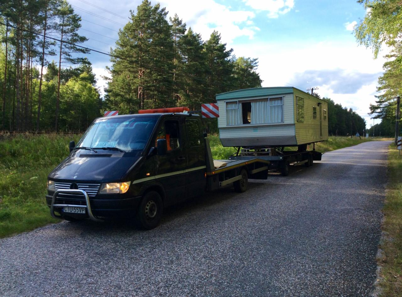 Czarna laweta z pomarańczowym światłem ostrzegawczym transportuje przyczepę kempingową po asfaltowej drodze w otoczeniu lasu i zieleni.