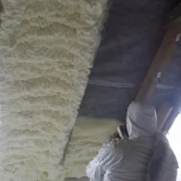 Ocieplenie poddasza pianką PUR: biała, nierówna struktura pianki pokrywa powierzchnię, widoczny pracownik w kombinezonie ochronnym podczas aplikacji.