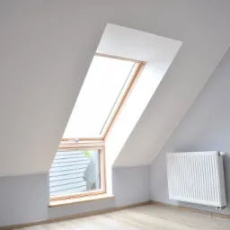 Poddasze z oknem dachowym w drewnianej ramie, pomalowane ściany w odcieniach szarości i biały grzejnik, widok na dach sąsiedniego budynku.
