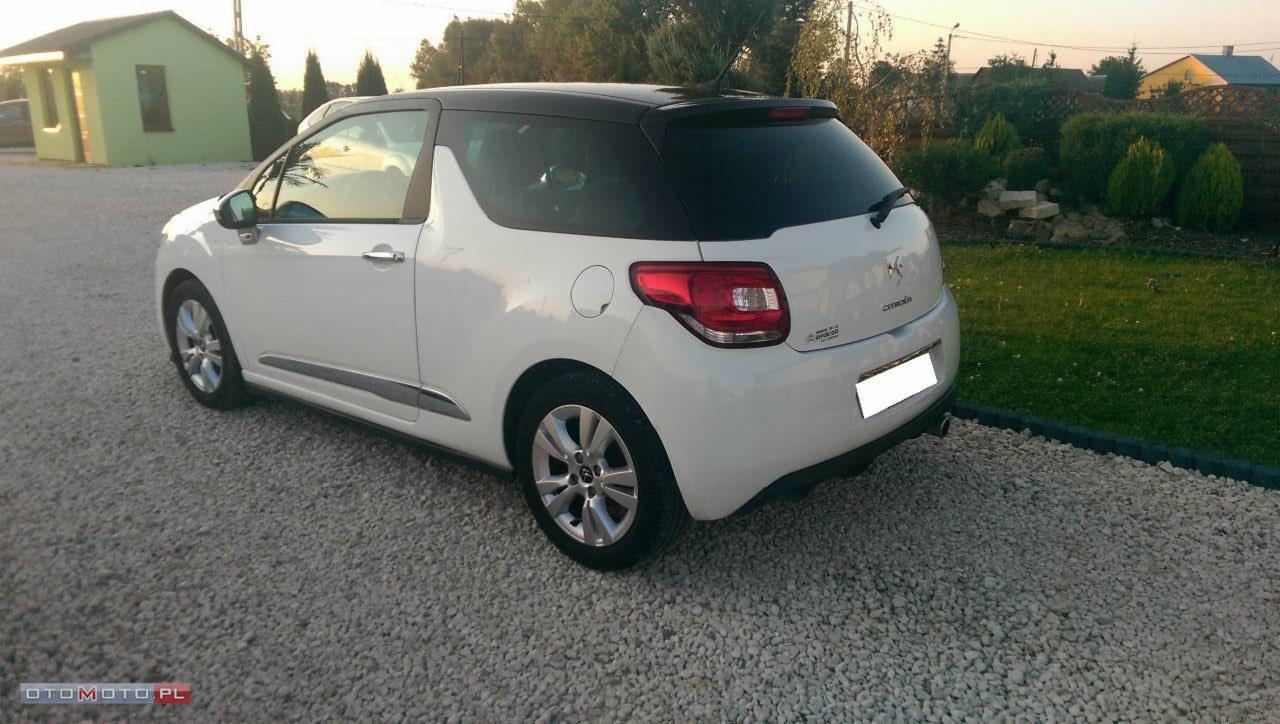 Biały Citroën DS3 z czarnym dachem, zaparkowany na żwirowym podjeździe przed zielonym budynkiem, widok z tyłu.