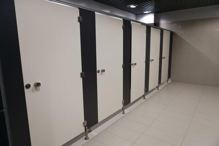 Rząd kabin sanitarnych z białymi drzwiami i ciemnymi panelami bocznymi, wspartych na stalowych nóżkach, widocznych na tle jasnej podłogi z płytek.