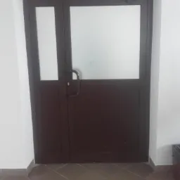 Ciemnobrązowe drzwi z matowym przeszkleniem, widoczny samozamykacz, kafelki na podłodze.