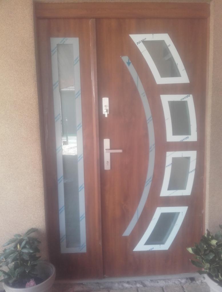 Brązowe drzwi wejściowe z przeszkleniami, zabezpieczone folią ochronną, z widocznym zamkiem i klamką, ustawione w ścianie obok doniczki z zieloną rośliną.