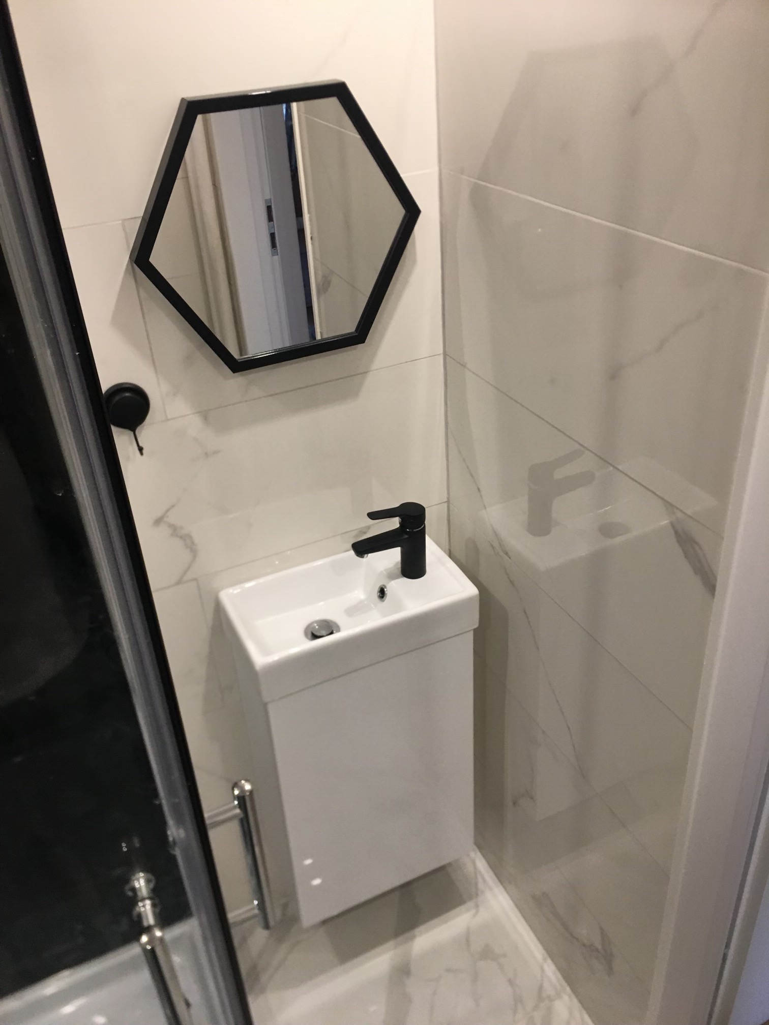 Mała, nowoczesna łazienka z białą umywalką podwieszaną, czarną baterią i heksagonalnym lustrem w czarnej ramie, wykończona płytkami imitującymi marmur.