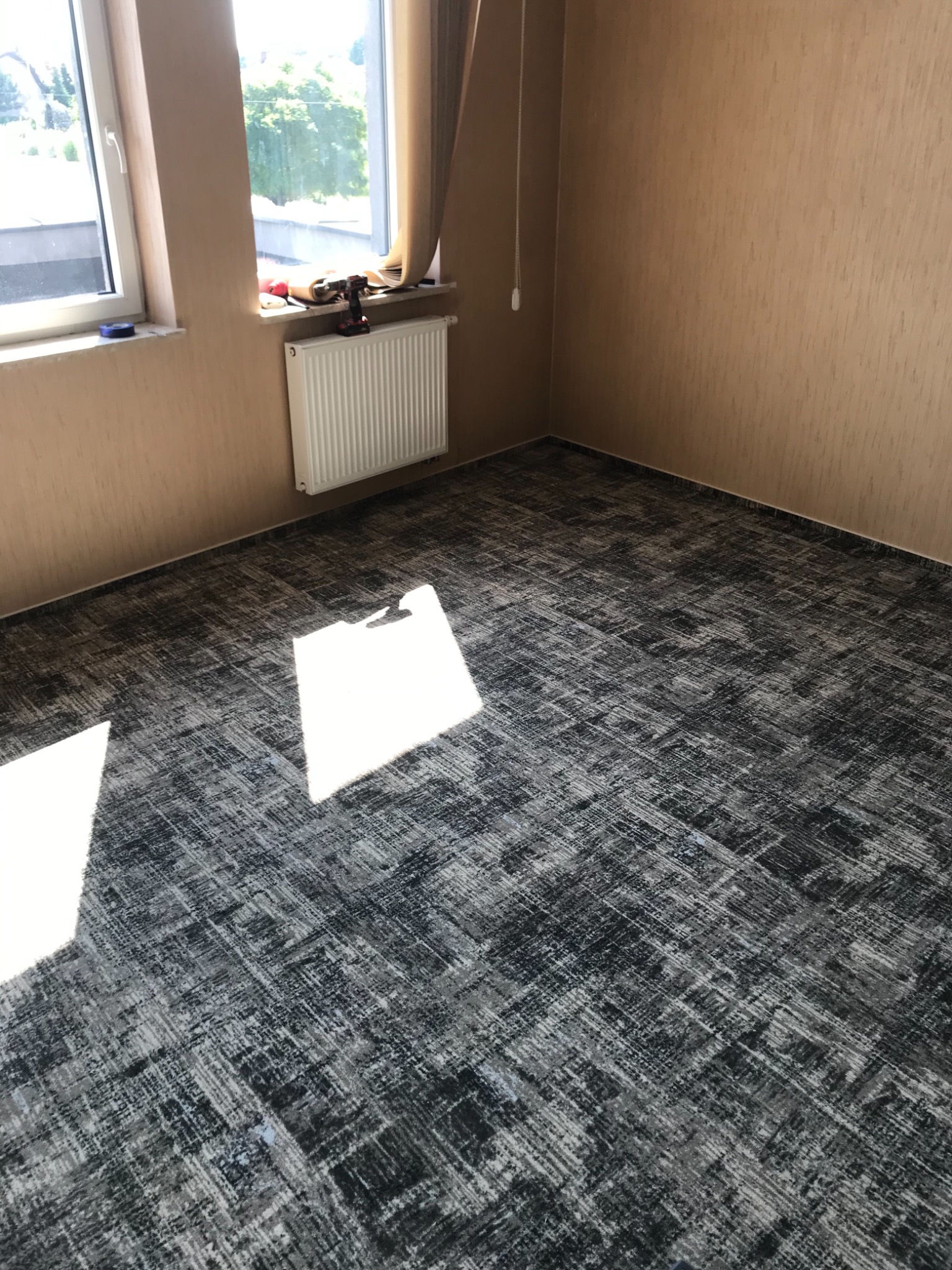 Pokój po remoncie z nową wykładziną w odcieniach szarości i beżu, widoczne narzędzia na parapecie, ściany w beżowej strukturze.