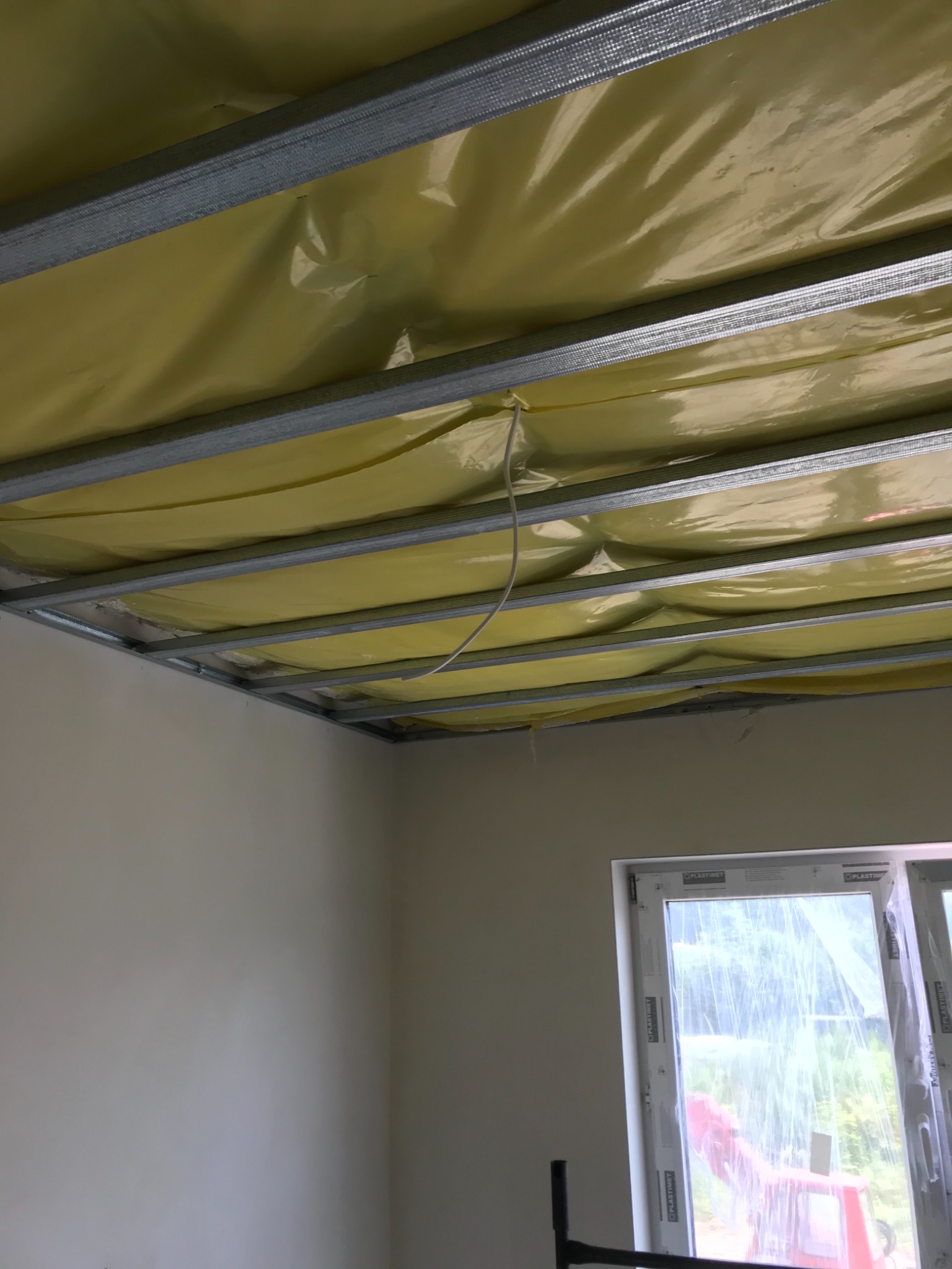 Montaż sufitu napinanego: metalowa konstrukcja podwieszana z żółtą folią, widoczny fragment okna zabezpieczonego folią.