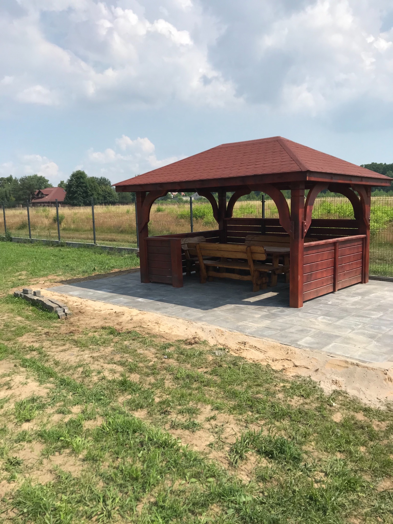 Nowo wybudowana, drewniana altana z czerwonym dachem, wyposażona w stół i ławki, umieszczona na wyłożonym kostką brukową tarasie, otoczona trawnikiem i ogrodzeniem z siatki.