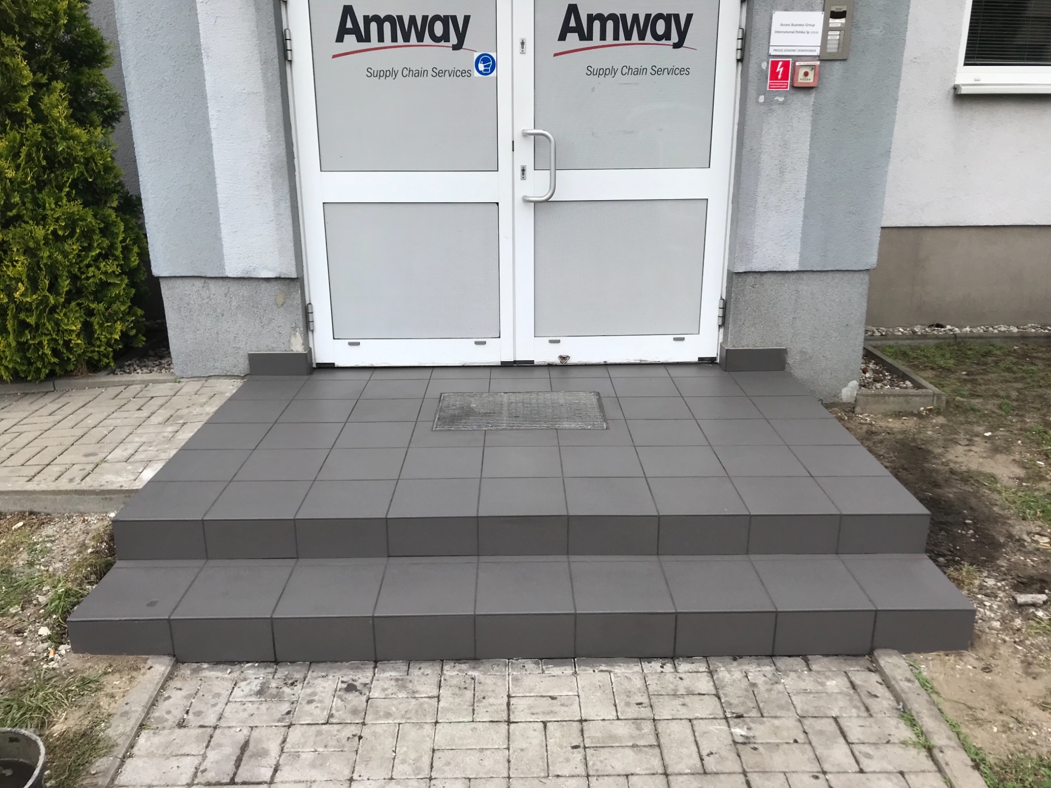 Wyłożone szarymi płytkami schody wejściowe do budynku Amway z widoczną kratką ściekową przed drzwiami.