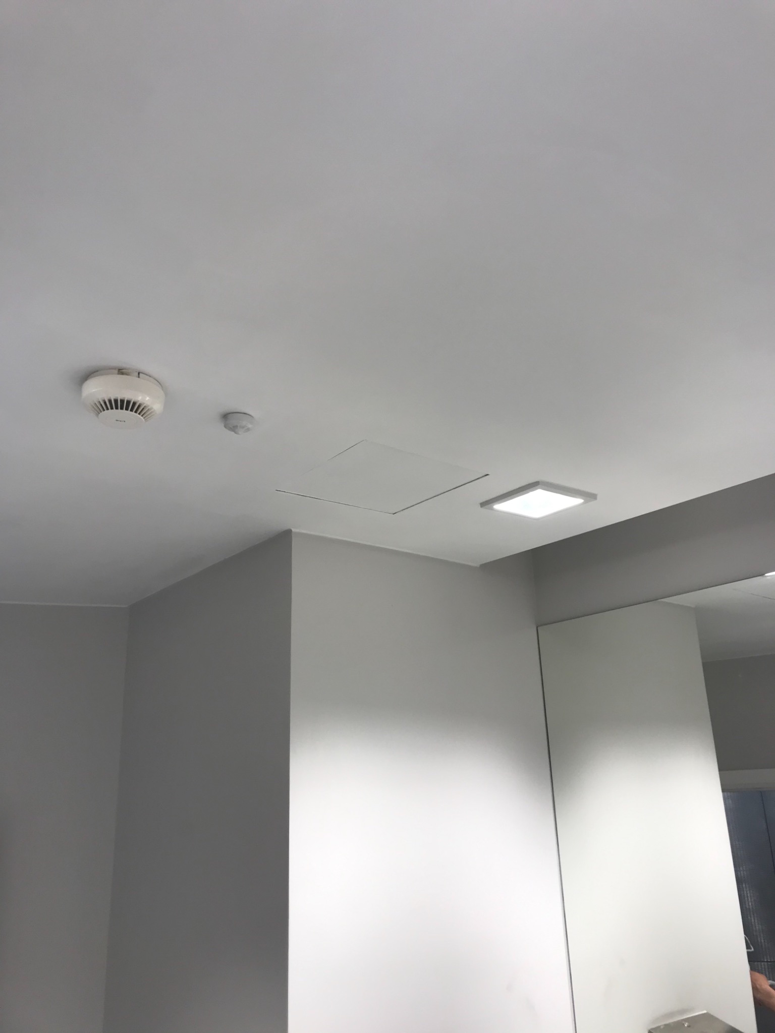 Minimalistyczna, świeżo pomalowana łazienka z białym sufitem, czujnikiem dymu, kwadratową lampą LED wpuszczoną w sufit i lustrem z szarymi ścianami.