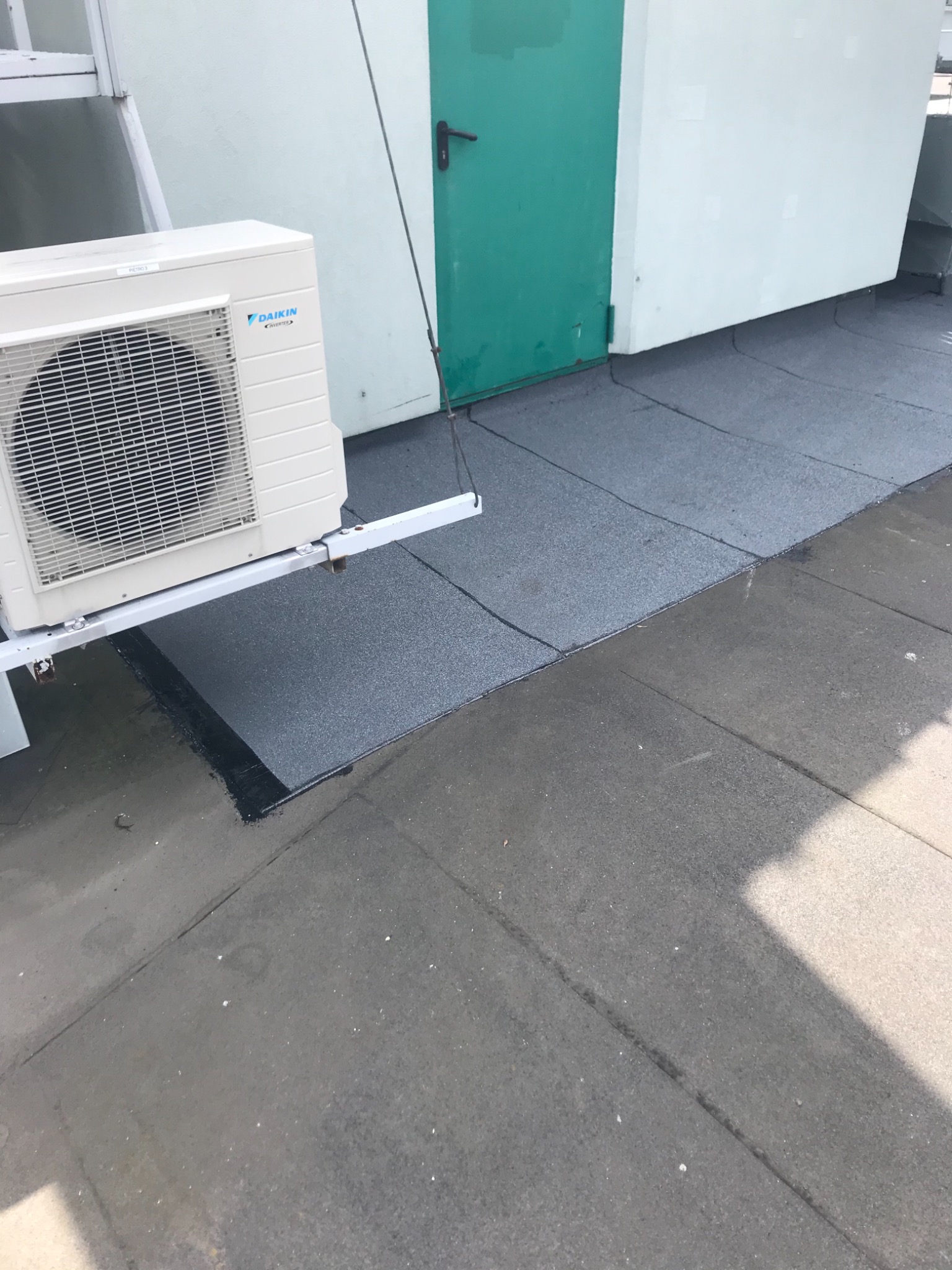 Fragment dachu pokrytego papą termozgrzewalną z widocznym agregatem klimatyzacyjnym Daikin na metalowym wsporniku oraz zielonymi drzwiami w tle.