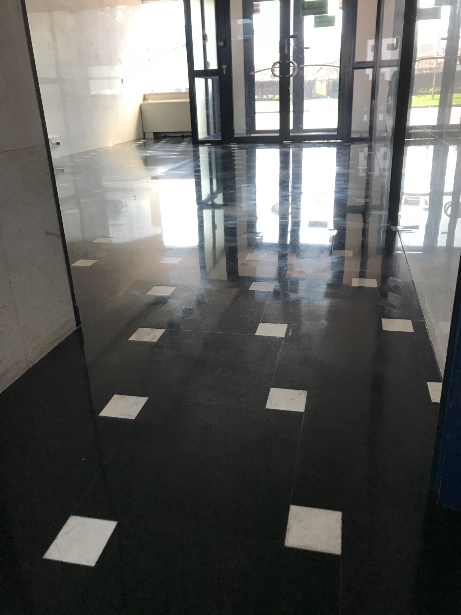 Błyszcząca, czarna podłoga z kwadratowymi, białymi wstawkami, odbijająca światło z przeszklonych drzwi wejściowych, widoczne ślady po remoncie na ścianach.