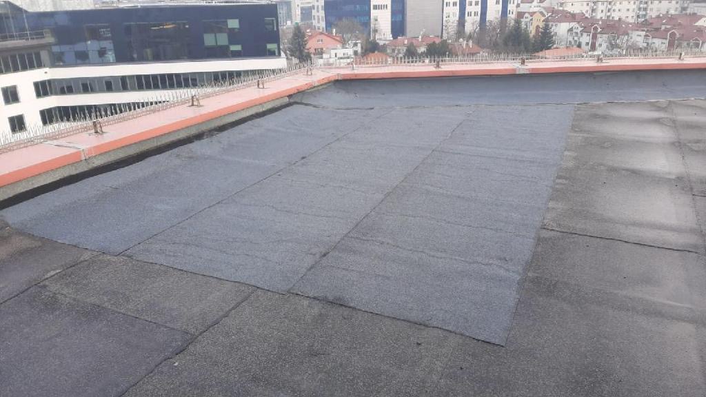Fragment płaskiego dachu budynku pokrytego nową papą termozgrzewalną, widoczne łączenia arkuszy, w tle budynki miejskie i zabezpieczenia przeciw ptakom.