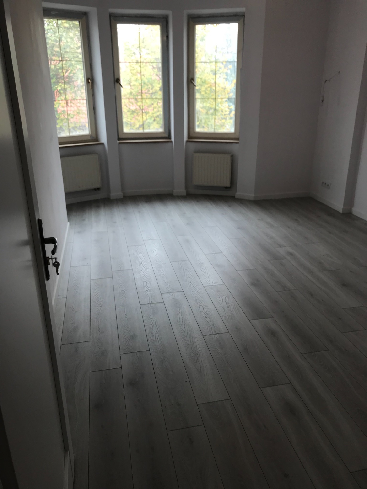 Pusty pokój po remoncie z panelami podłogowymi w odcieniach szarości i trzema oknami z widokiem na drzewa, widoczny uchwyt drzwi z kluczem.