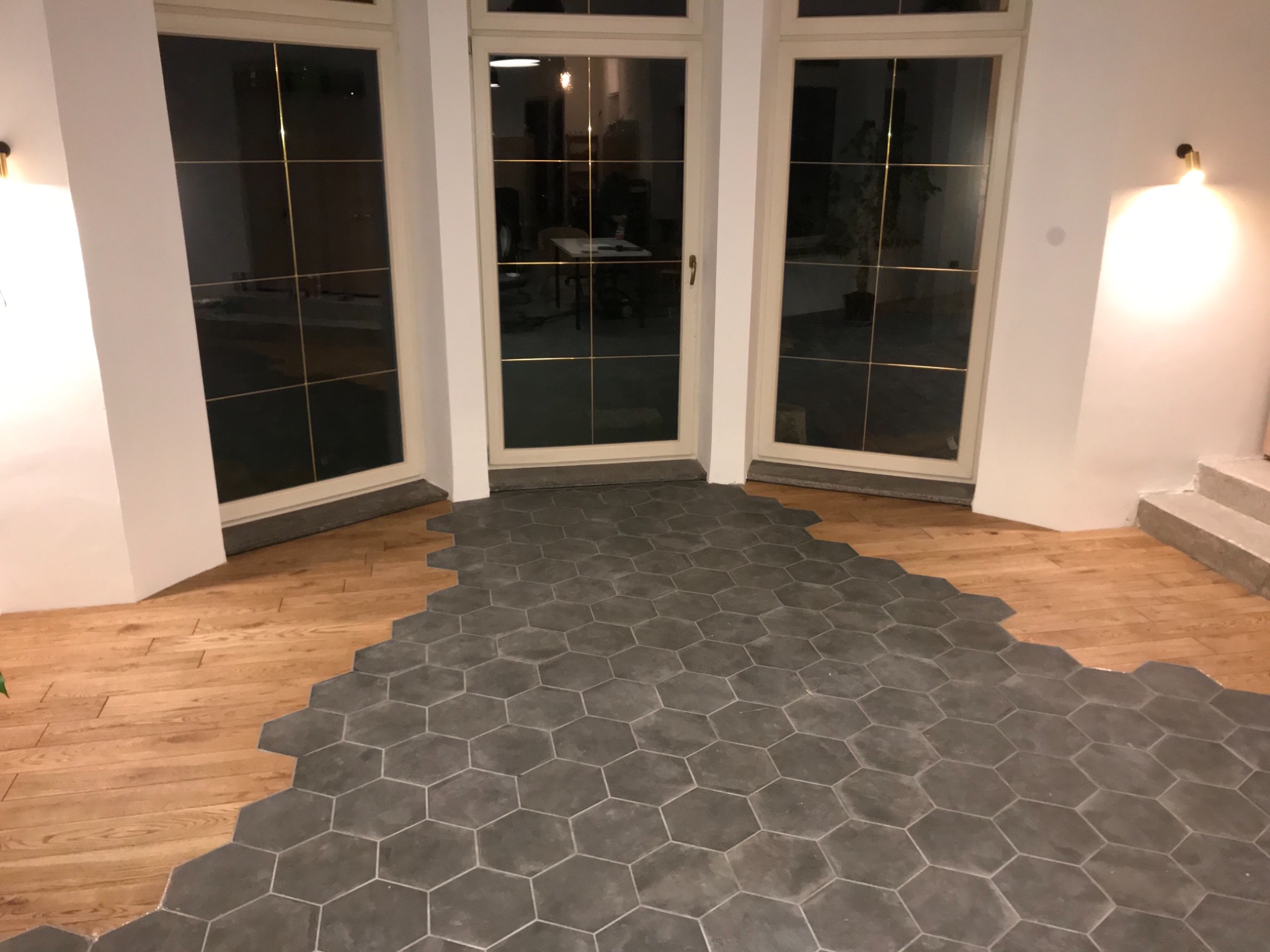 Układanie heksagonalnych, grafitowych płytek podłogowych w połączeniu z drewnianą podłogą w jasnym wnętrzu z trzema oknami.