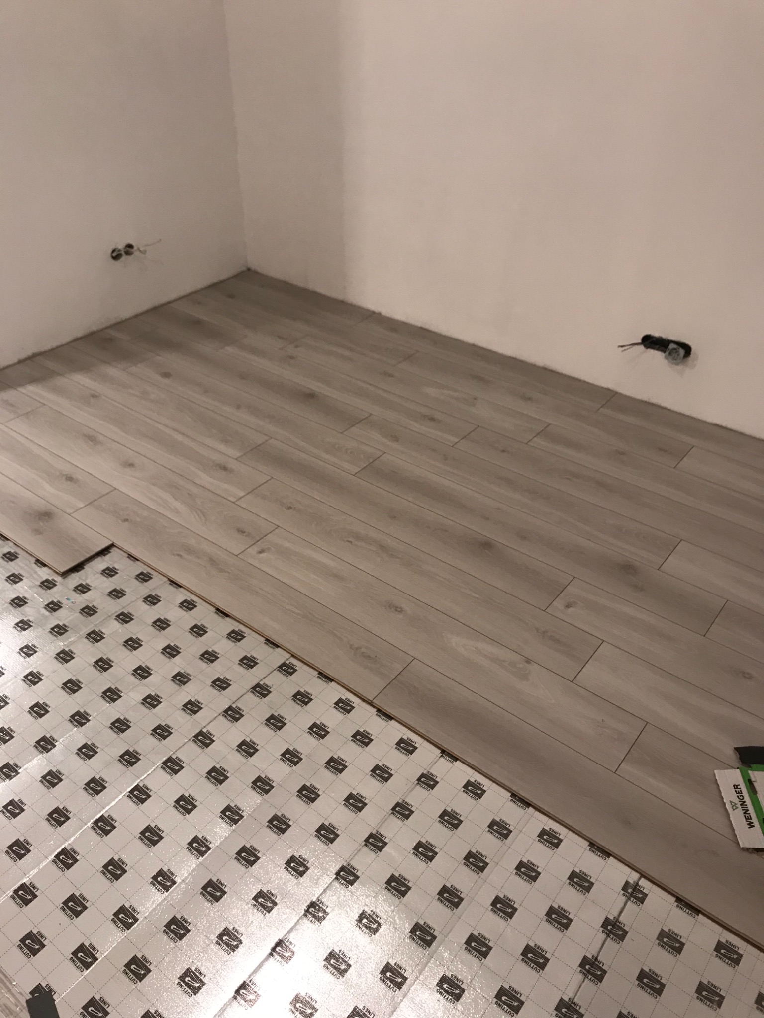 Układanie paneli podłogowych na podkładzie izolacyjnym w rogu pomieszczenia z widocznymi gniazdkami elektrycznymi.