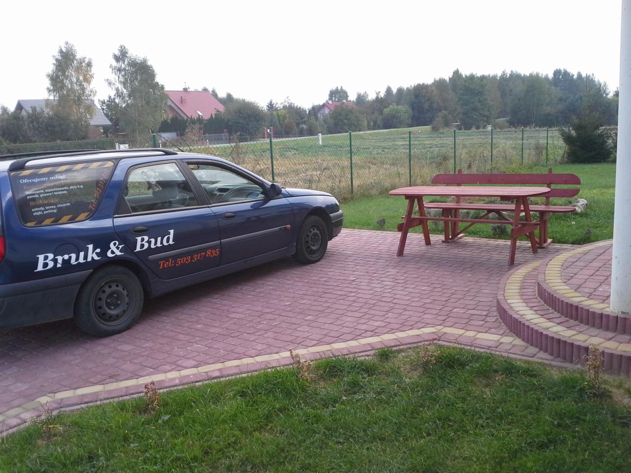 Niebieski samochód firmy brukarskiej 'Bruk & Bud' zaparkowany na wybrukowanym podjeździe z czerwonej kostki, obok drewnianego stołu z ławkami na tle zieleni.