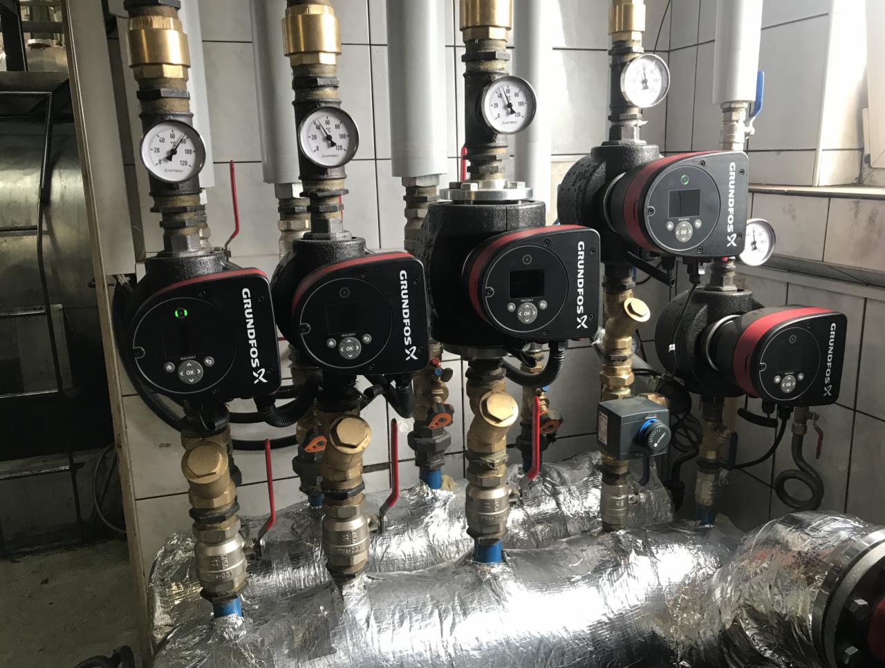 Rząd pomp obiegowych Grundfos z manometrami, zamontowanych na rurach owiniętych folią izolacyjną, na tle białych płytek.