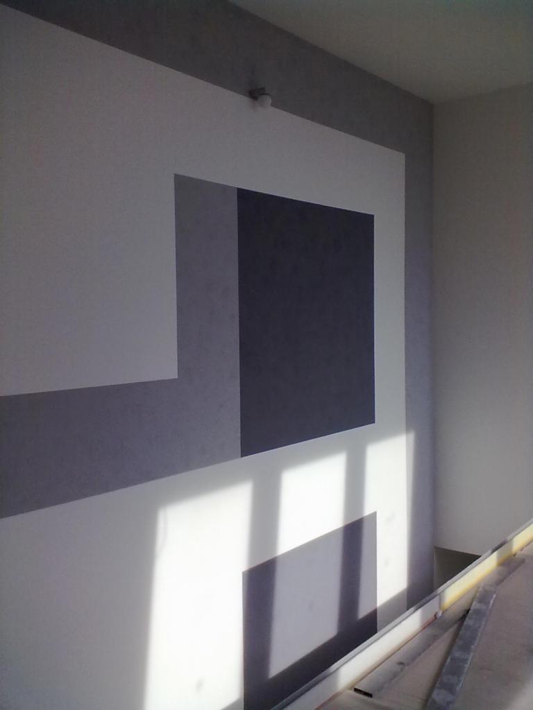 Geometryczny wzór na ścianie w odcieniach szarości i bieli, oświetlony naturalnym światłem. Nowoczesny design z widocznymi elementami wykończeniowymi w dolnej części kadru.