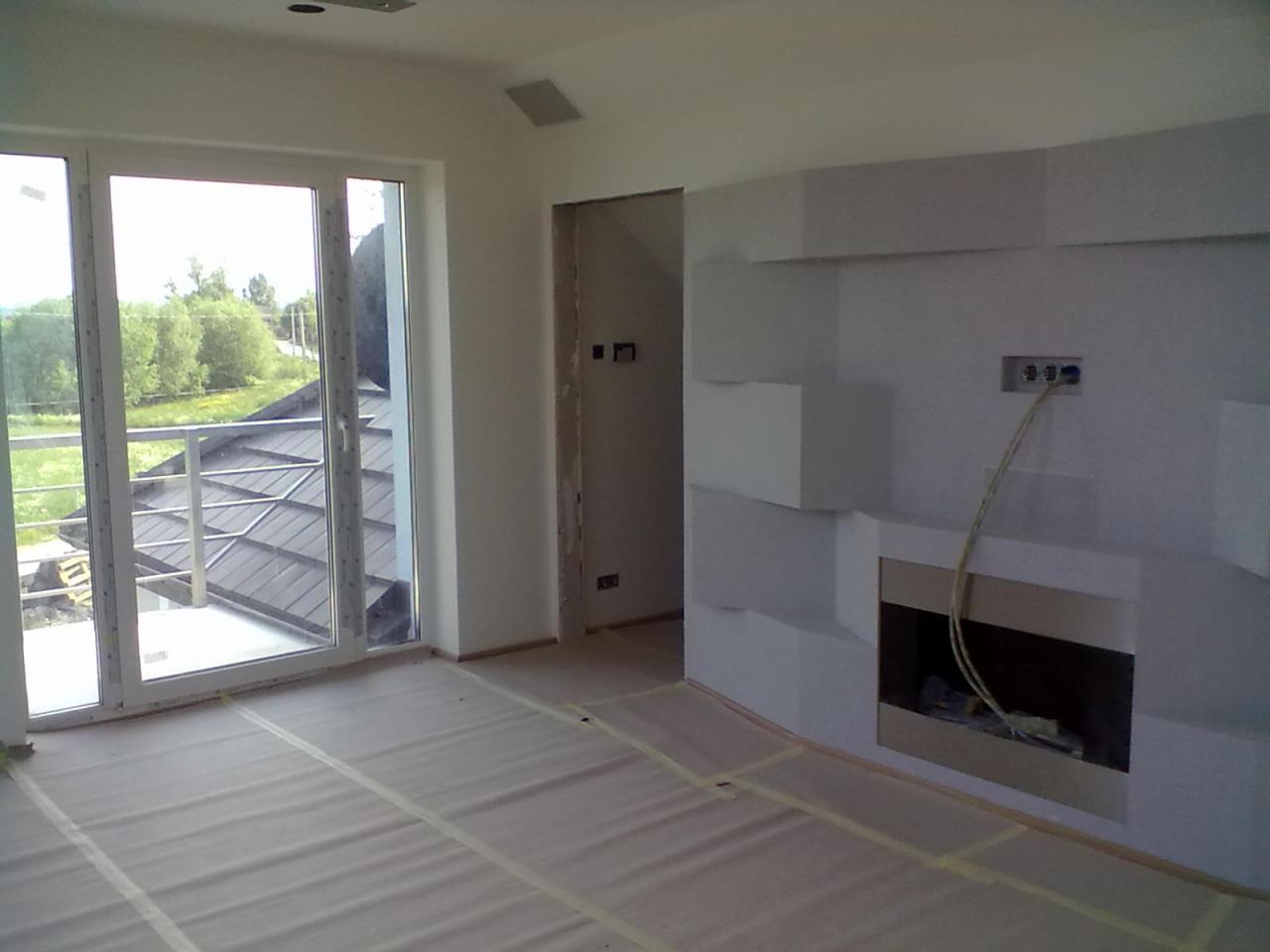 Wnętrze w trakcie wykańczania: zabezpieczona podłoga, nowoczesny kominek z dekoracyjną ścianą, duże okno balkonowe z widokiem na zewnątrz.