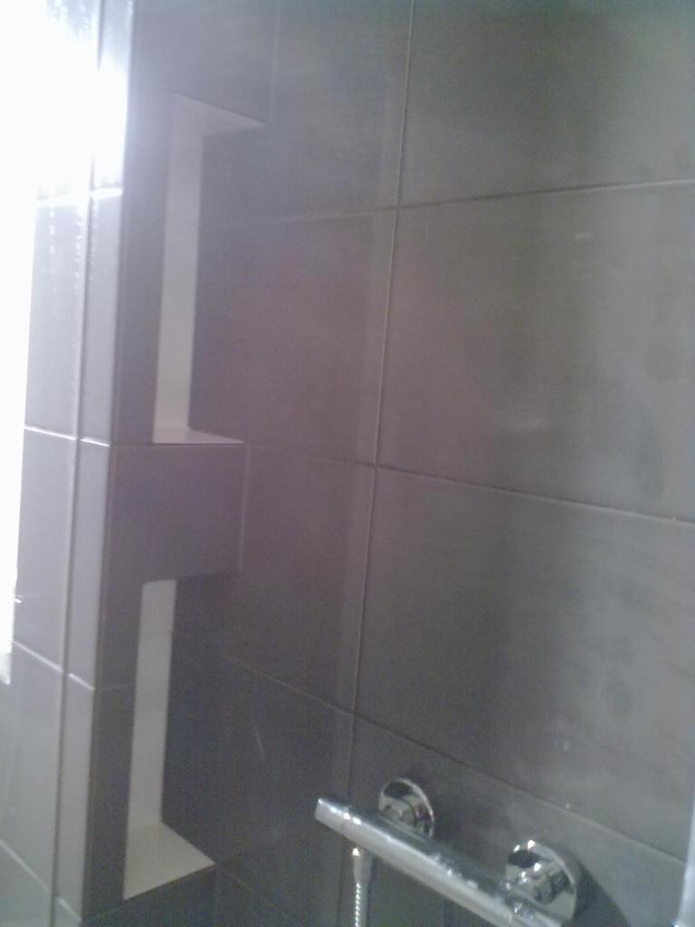 Szare płytki w łazience z wnęką i chromowaną armaturą. Nowoczesne wykończenie ściany w kabinie prysznicowej, widoczny fragment baterii.