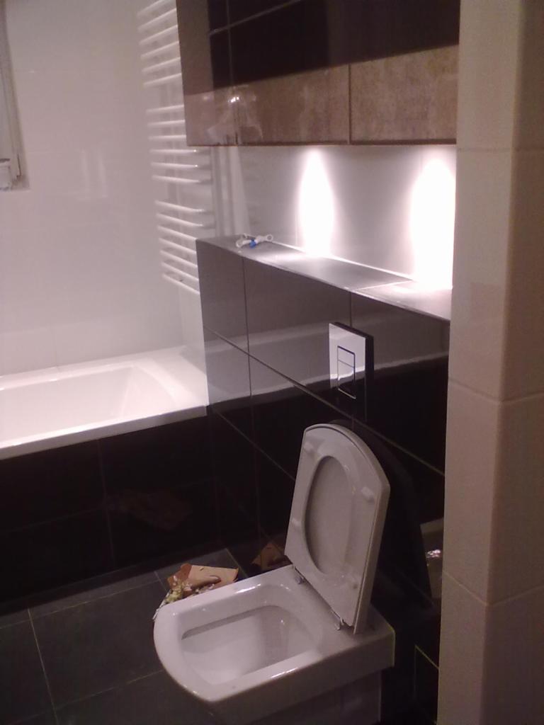 Nowoczesna łazienka z czarnymi płytkami, podświetlanymi szafkami i białą wanną. Uwagę przyciąga otwarta klapa sedesu.