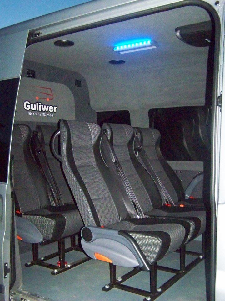 Wnętrze busa z szarymi, tapicerowanymi fotelami z pasami bezpieczeństwa, oświetlone niebieską lampą LED. Widoczne logo 'Guliwer Express Europa'.