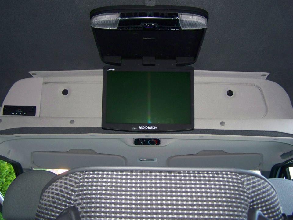 Wnętrze busa z zamontowanym ekranem Audiomedia na suficie, oświetleniem i panelem sterowania. Widoczne fotele z wzorem w kratkę i szara tapicerka.