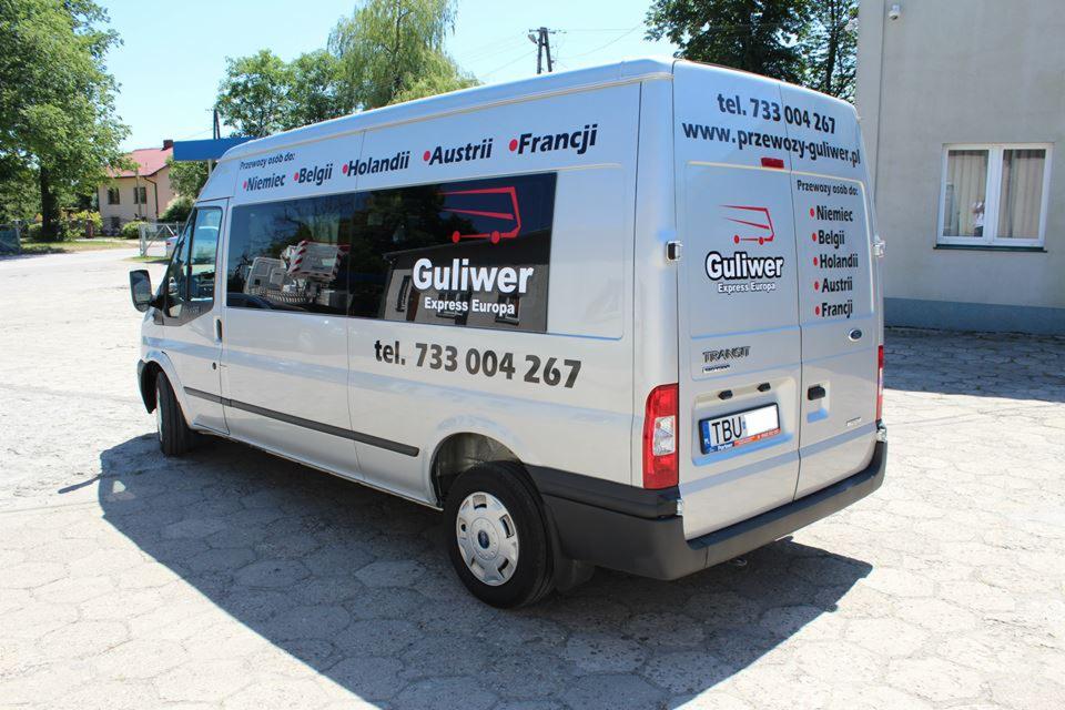 Srebrny bus z logo firmy przewozowej 'Guliwer Express Europa' i numerem telefonu, zaparkowany na zewnątrz budynku. Widoczne kraje docelowe: Niemcy, Belgia, Holandia, Austria, Francja.