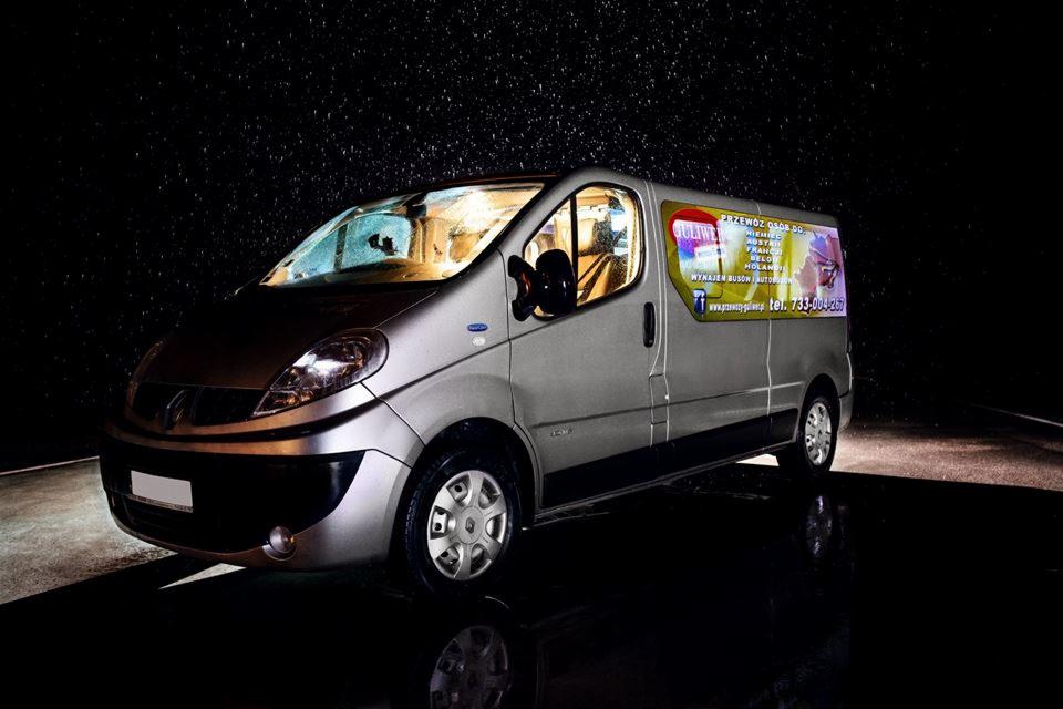 Szary bus Renault Trafic z reklamą przewozu osób, oświetlony w nocy. Widoczna reklama z numerem telefonu i kierunkami przewozów.