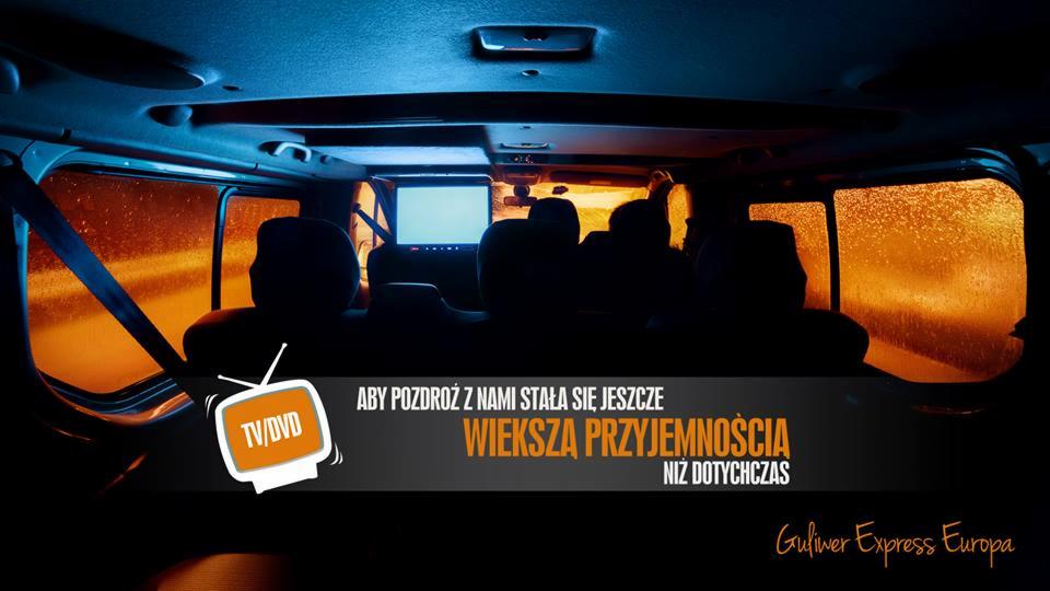 Wnętrze busa z ekranem DVD, widok z tyłu na siedzenia pasażerów. Za oknem deszcz. Reklama firmy przewozowej z hasłem o przyjemności podróży.