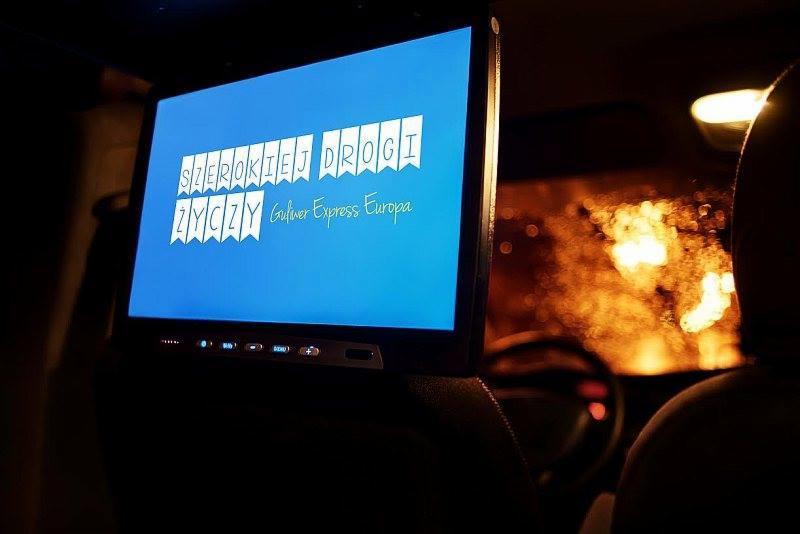 Ekran z życzeniami 'Szerokiej Drogi' w ciemnym wnętrzu pojazdu, prawdopodobnie podczas przewozu osób. Widoczne siedzenia i deska rozdzielcza.