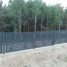 Nowoczesne, poziome, szare przęsła ogrodzeniowe na betonowej podmurówce, w tle las mieszany.