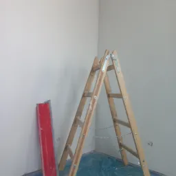 Świeżo wykończone, białe ściany w rogu pokoju zabezpieczone folią, drewniana drabina rozstawiona, obok ściany oparta czerwona szpachelka.
