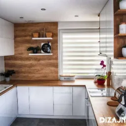 Nowoczesna kuchnia z białymi, lakierowanymi szafkami, drewnianym blatem i panelem ściennym, oświetleniem punktowym oraz półkami z dekoracjami i roślinami doniczkowymi.