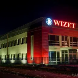 Oświetlony nocą budynek firmy Wizet Polska, z czerwono-białą elewacją i dużym logo na dachu, widoczny z ulicy za metalowym ogrodzeniem.