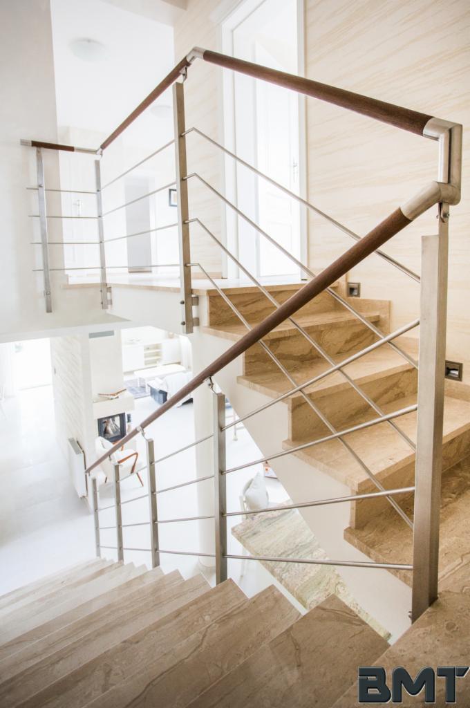 Marmurowe schody z metalową balustradą i drewnianym poręczą, widok z góry, jasne wnętrze, minimalistyczny design.