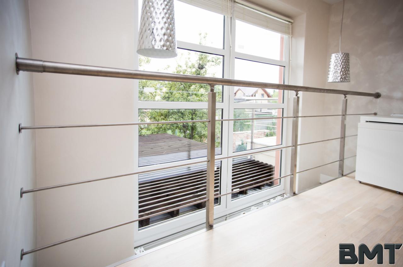 Nowoczesna balustrada ze stali nierdzewnej, z widokiem na zewnątrz przez duże okno. Minimalistyczny design, jasne wnętrze, wiszące lampy dekoracyjne.