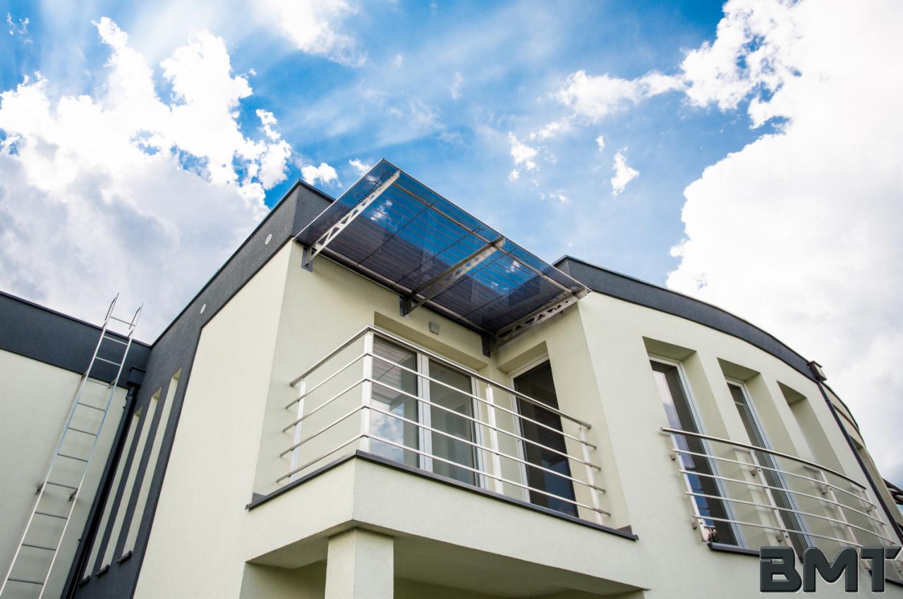 Nowoczesny dom z balkonem z chromowaną balustradą i zadaszeniem z poliwęglanu, widziany z dołu na tle błękitnego nieba z chmurami.