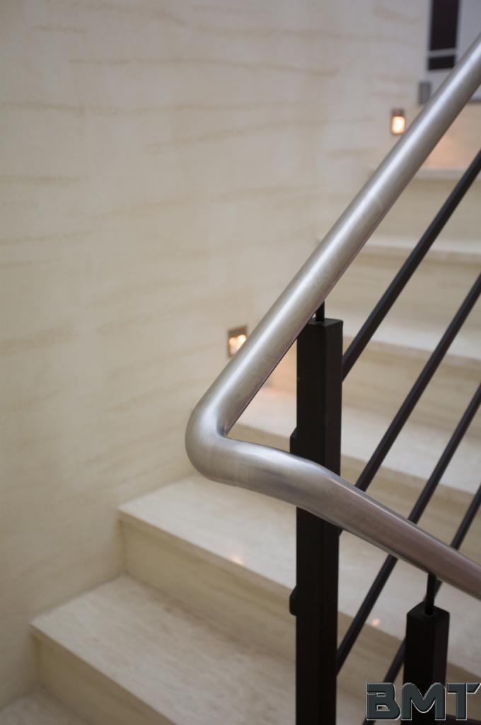 Minimalistyczna balustrada schodowa z metalową poręczą i czarnymi słupkami, oświetlona punktowymi lampami w ścianie, na tle marmurowych schodów.
