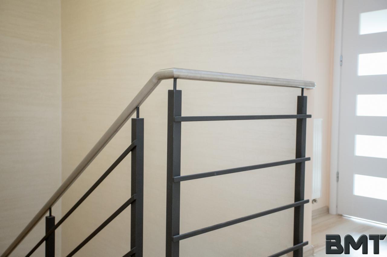 Minimalistyczna balustrada metalowa z poziomymi prętami i drewnianą poręczą, widoczna na tle kremowej ściany i białych drzwi w nowoczesnym wnętrzu.