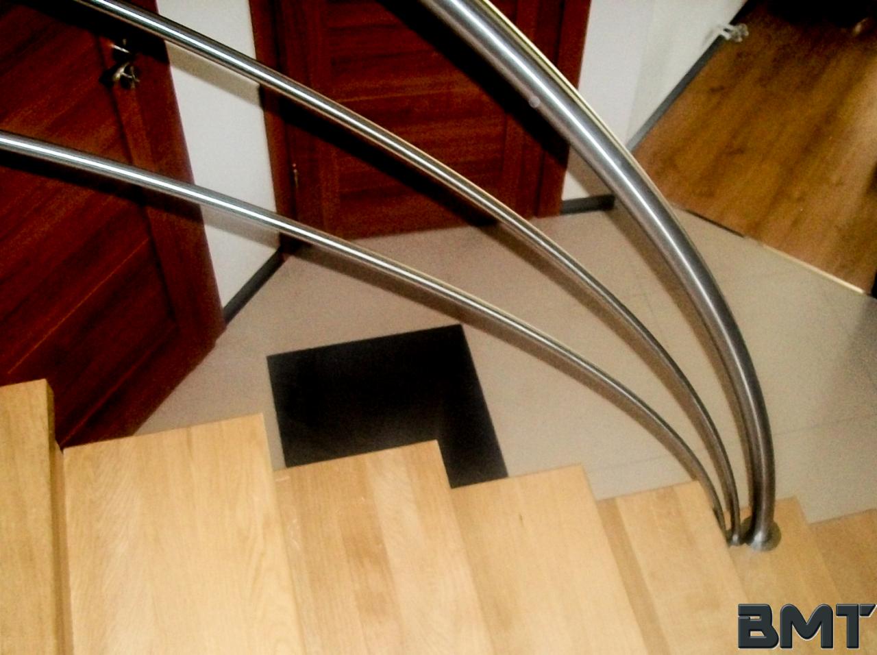 Drewniane schody z metalową, giętą balustradą w nowoczesnym wnętrzu, widok z góry na pierwsze stopnie. W tle widoczne drzwi i fragment podłogi.