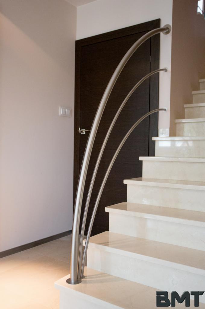 Nowoczesna balustrada metalowa z trzema łukowatymi prętami na marmurowych schodach, z ciemnymi drzwiami w tle i logo BMT w prawym dolnym rogu.