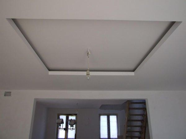 Biały sufit z dekoracyjnym wgłębieniem w kształcie kwadratu, z widoczną żarówką na kablu. W tle widoczne schody i okna, wnętrze w trakcie wykańczania.