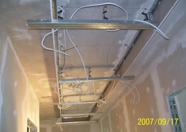 Sufit podwieszany w trakcie montażu, widoczne profile metalowe, kable, szpachlowane ściany. Ujęcie z dołu, perspektywa wnętrza w trakcie prac wykończeniowych.