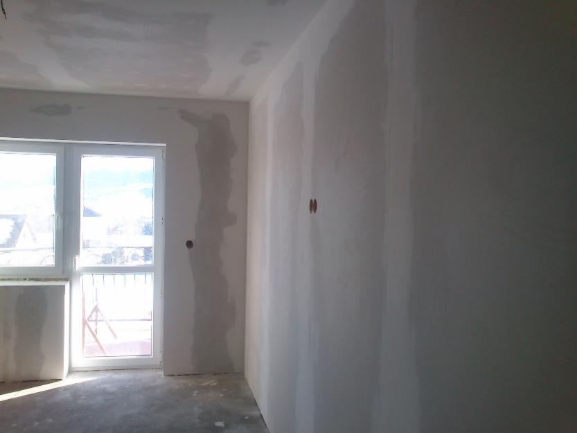 Pokój po szpachlowaniu: gładkie ściany przygotowane do malowania, widoczne ślady po łataniu, okno z widokiem na zewnątrz. Przygotowanie do dalszych prac wykończeniowych.