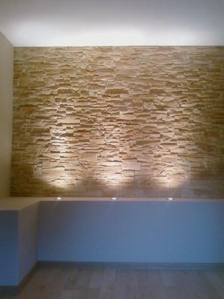 Ściana wykończona kamieniem dekoracyjnym w odcieniach beżu, oświetlona punktowymi lampami, tworząc ciepły i nowoczesny efekt wizualny we wnętrzu.